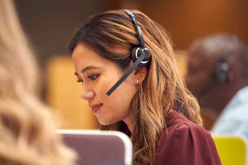 Mature Businesswoman Wearing Phone Headset Talking To Caller In Busy Customer Services Centre